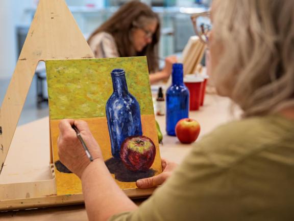 Adult student paints still life of bottle and apple on table top easel (SAC)